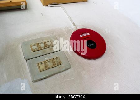 This image features a red emergency button set against a white wall, accompanied by beige light switches, highlighting safety and alert systems in des Stock Photo