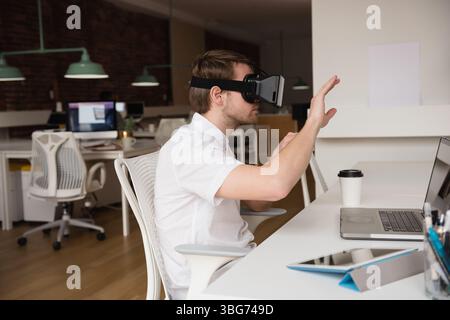 Male office worker wearing VR headset sitting at desk in open-plan office raising hand with laptop Stock Photo