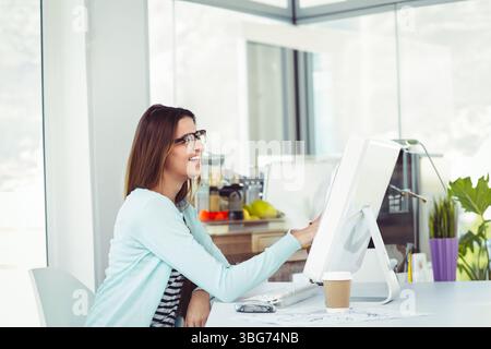 All-in-one computer glowing on desk, sketches resting and coffee steaming in glass-walled office Stock Photo