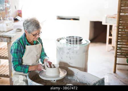Senior female potter in apron shaping clay vessel on pottery wheel in ceramics studio, copy space Stock Photo