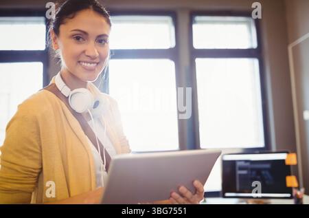 Hispanic woman holding tablet with headphones near desk monitor and orange notes at office windows Stock Photo