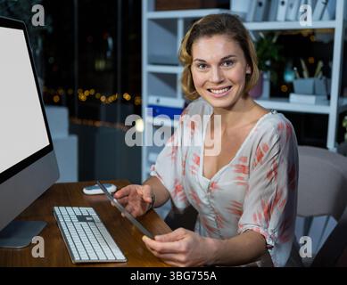 Woman in coral blouse holding tablet and using monitor on wooden desk at night, copy space Stock Photo