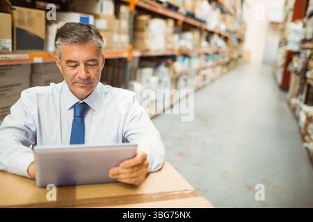 Man in shirt and tie checking inventory on tablet leaning on boxes in warehouse, copy space Stock Photo