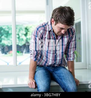 Man sitting on wide white window ledge, leaning forward in bright modern room with garden view Stock Photo