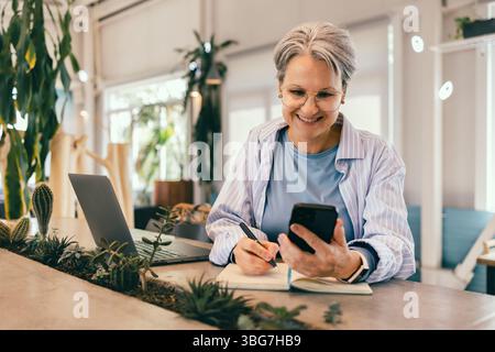 Smiling senior woman with gray hair writing notes while using smartphone at cozy modern workspace Stock Photo