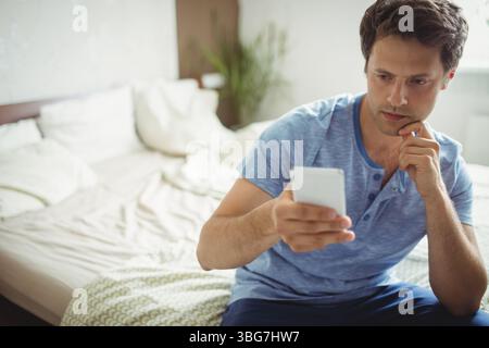 Middle-aged man checking smartphone while resting chin on hand on bed with plant, copy space Stock Photo
