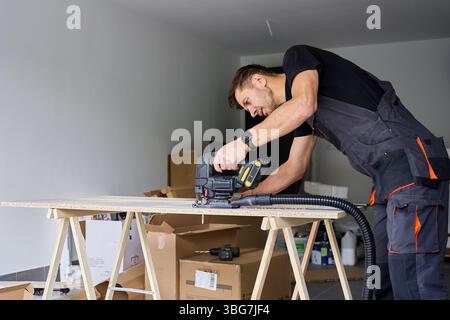 Worker in workwear using handheld cordless electric jigsaw to cut wooden board in garage. Skilled carpenter working in workshop. Man doing DIY home re Stock Photo