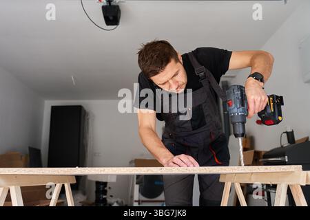 Man in work overalls bib using cordless power drill to make hole in laminated wood board. Male carpenter working in home workshop, drilling chipboard Stock Photo