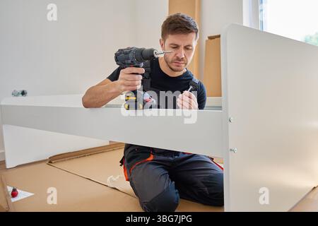 Focused man in bib overalls connects details of new bed in bedroom using electric drill. Assembling furniture at home Stock Photo