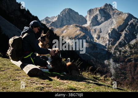 Smiling woman sitting with two dogs on a bench in the Komovi mountains of Montenegro, enjoying a sunny autumn day with stunning rocky peaks in the Stock Photo