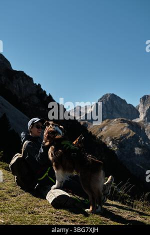 Smiling woman sitting with two dogs on a bench in the Komovi mountains of Montenegro, enjoying a sunny autumn day with stunning rocky peaks in the Stock Photo