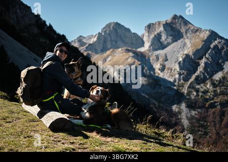 Smiling woman sitting with two dogs on a bench in the Komovi mountains of Montenegro, enjoying a sunny autumn day with stunning rocky peaks in the Stock Photo
