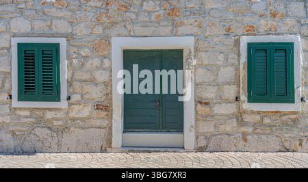 Symmetrical stone wall facade with a green double door in the center and two green shuttered windows on either side Stock Photo