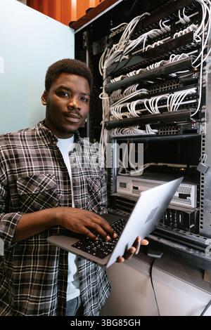 Man working on a laptop in a server room during daylight hours, managing network equipment and data connections Stock Photo