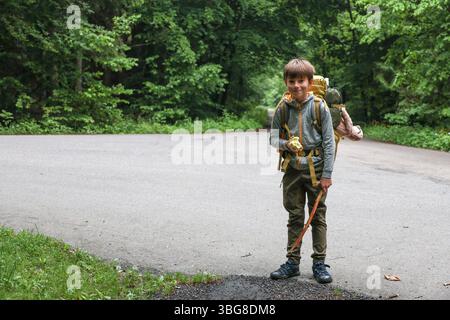 Smiling boy with backpack hiking in forest, holding apple and walking stick, outdoor childhood adventure Stock Photo