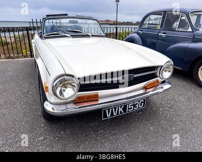 Front view of a classic white TR6 convertible car parked on a paved area beside an iconic vintage car under a cloudy sky Stock Photo