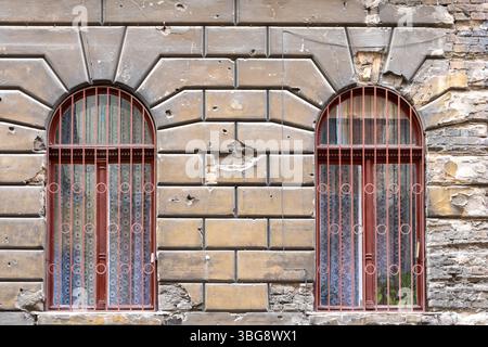 Textured background of old brickwork with bullet and shell marks from the war and two arched windows with bars Stock Photo