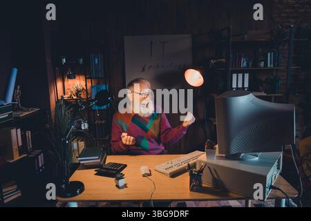 Elderly male programmer working on computer in home office illuminated by warm light Stock Photo