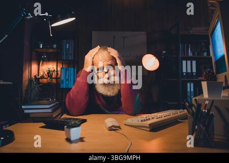Elderly man working late on computer in cozy home office setting with focused expression, showcasing professional dedication Stock Photo