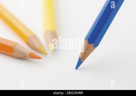 Flat-angled detail view of a blue wooden coloured pencil placed diagonally on paper in front of three flat coloured pencils in shades of yellow, the t Stock Photo