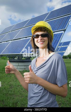 Upper body view of a young woman with construction site helmet and sunglasses in front of a photovoltaic plant pointing both thumbs up and smiling int Stock Photo