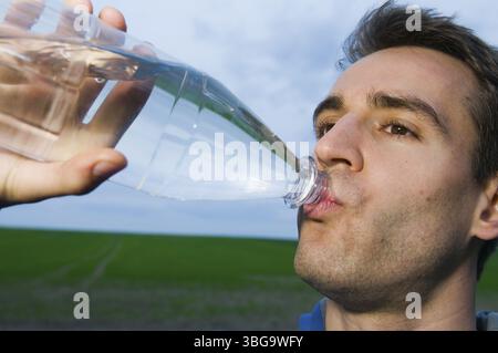 Side head-and-shoulders shot of a young man in a blue T-shirt and blue towel around his neck drinking from a plastic bottle in front of a green meadow Stock Photo
