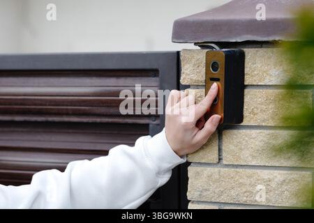 Woman using smart video intercom system, pressing button on electronic device installed on brick wall near entrance gate Stock Photo