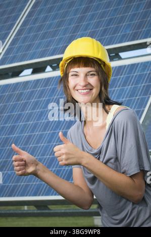 Upper body view of a young brunette woman in a yellow construction helmet standing sideways in front of a photovoltaic system, pointing both thumbs up Stock Photo