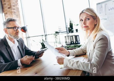 Mature businesspeople collaborating in a modern office setting, discussing documents and strategies at a wooden desk Stock Photo
