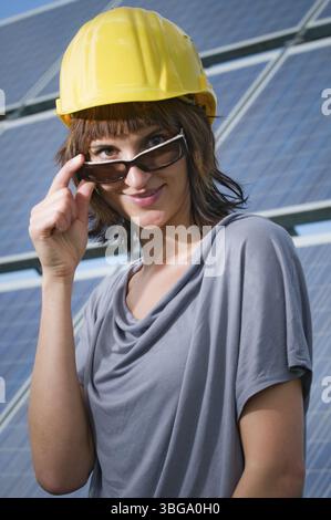 Upper body shot of a young, brunette woman in a yellow construction helmet looking over sunglasses held with her right hand towards the camera, backgr Stock Photo