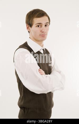 Upper body shot of a young man in a white shirt and brown vest over a white background with folded arms looking half sideways into the camera without Stock Photo