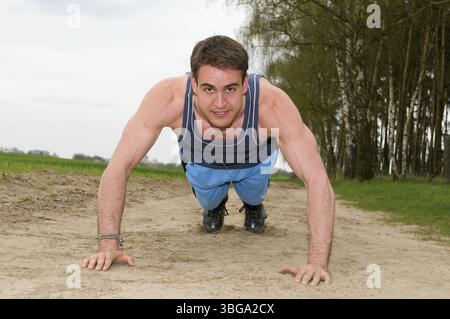 Frontal full-body shot of a man facing the camera in short blue sportswear on a sandy path in a push-up position looking into the camera Stock Photo