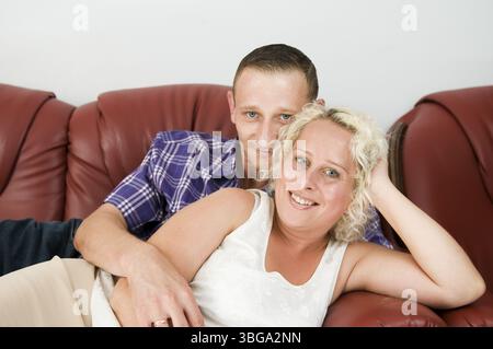 Upper body portrait of a young couple lying one behind the other on a brown leather sofa and smiling happily into the camera Stock Photo