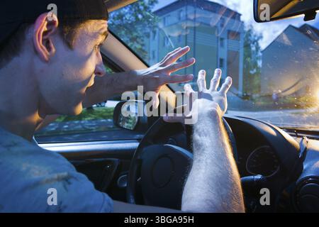 Side wide-angle upper body interior view of a blinded car driver (young man) holding his hands protectively in front of him towards the windscreen wit Stock Photo