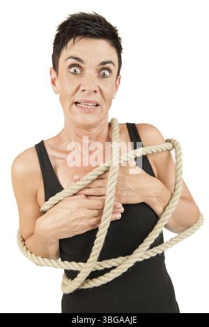 Frontal upper body view of a black-haired middle-aged woman in a black top with a rope around her body looking shocked into the camera in front of a w Stock Photo
