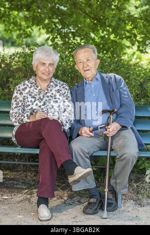 Full-length portrait of an 80-year-old pensioner couple on a park bench in the shade of a canopy of leaves in summer, looking joyfully into the camera Stock Photo