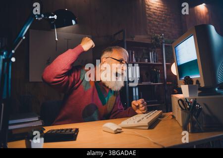 Enthusiastic elderly technology specialist celebrates achievement while working late at home office during an evening programming session Stock Photo