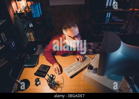 Elderly man working on computer in a cozy office space with focus on technology and creativity during evening hours Stock Photo