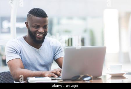 Smiling african man textmessaging on laptop in cafe Stock Photo