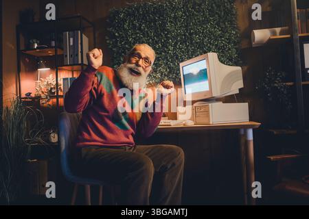 Cheerful elderly man wearing a colorful sweater working on an old desktop computer in a cozy home office during the evening. Stock Photo