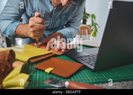 Leather craftsman working with leather using a stitching groover and laptop in his workshop Stock Photo