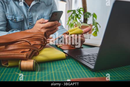 Leather craftsman working with genuine leather making handcrafted bags and wallets in a creative studio using smartphone and laptop Stock Photo