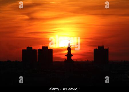 Dramatic sunset behind a city skyline featuring a silhouetted church tower and high-rise buildings under vivid orange and red skies. Stock Photo
