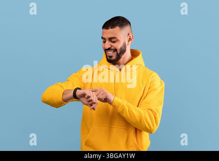 Young arab guy checking his smartwatch on blue studio background. Happy middle eastern man using fitness tracker Stock Photo