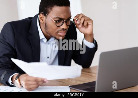 Black guy with glasses accountant holding papers, using laptop Stock Photo