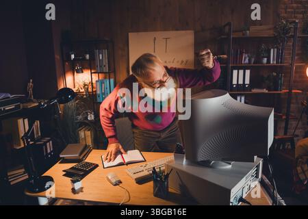 Elderly man in casual sweater frustrated with computer technology working in cozy home office interior during the evening Stock Photo