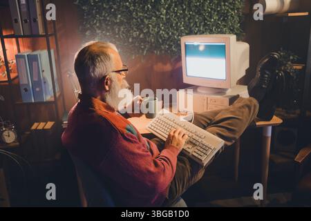 Mature man with white beard working on computer and drinking coffee in cozy home office with retro aesthetics Stock Photo