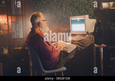 Elderly man working remotely on a vintage computer in a cozy home office surrounded by books and greenery Stock Photo