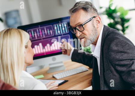 Experienced businessman discussing data with a colleague, analyzing statistics, and reviewing charts on a computer Stock Photo