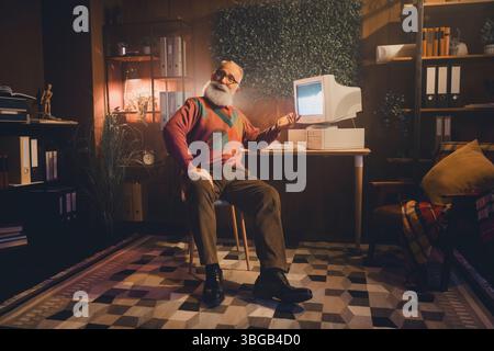 An elderly man seated at a desk, working with a retro computer in a cozy study space adorned with warm decor and light. Stock Photo